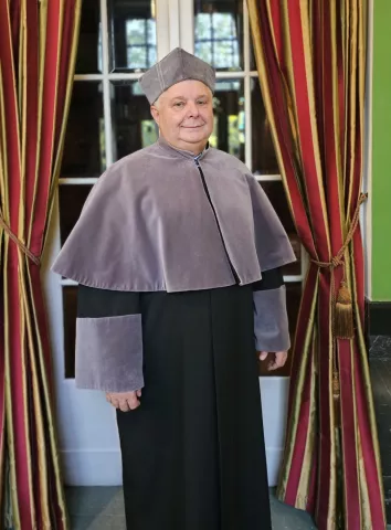 prodziekan w todze akademickiej na tle okna  z zasłonami
